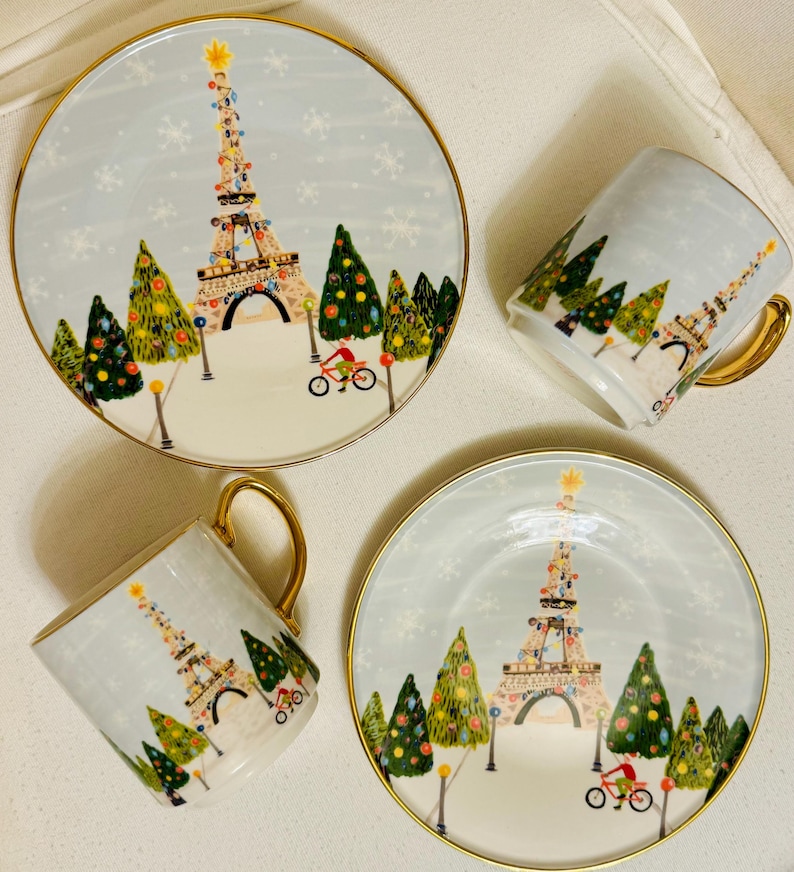 May include: A set of two plates and two mugs with a winter scene featuring the Eiffel Tower decorated with lights, Christmas trees, and a cyclist. The plates and mugs have a gold rim and handle.