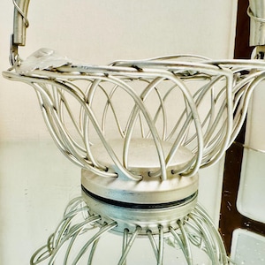 May include: A silver-toned metal basket featuring a wire-frame construction with a rounded base and a curved handle. The structure consists of multiple thin metal rods that form an open, lattice-like pattern, creating a bowl-shaped container.
