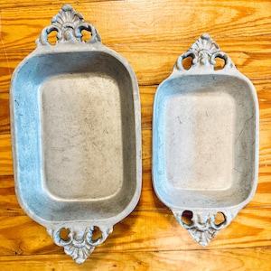 May include: Two rectangular metal trays with a matte gray finish and weathered texture. Each tray features ornate, decorative handles on both ends with a shell-like motif. The trays are positioned side by side, showing a larger and a smaller version.