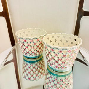 May include: Two cream-colored ceramic planters featuring a lattice cutout design. The exterior is decorated with a repeating pattern of small pink roses and green leaves, accented by thin blue horizontal bands near the scalloped top and base.