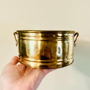 May include: A polished brass bowl with a cylindrical shape, featuring two small curved handles on opposite sides and horizontal ribbed detailing along the exterior surface. The metal has a reflective gold-toned finish with subtle surface variations.