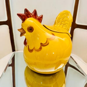 May include: A bright yellow ceramic chicken-shaped jar with a removable lid. The chicken features a dark red comb and wattle, brown painted eye details, and a textured tail feather design. The glossy finish reflects light across the rounded body.