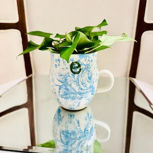 Peut inclure: Une tasse en céramique avec un motif floral bleu clair sur fond blanc et une lettre e minuscule vert foncé imprimée sur le devant. La tasse contient une plante aux feuilles vertes et possède une anse unique sur le côté droit.