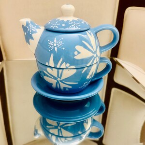 Może przedstawiać: Niebiesko-biały ceramiczny zestaw do herbaty z czajniczkiem, filiżanką i spodkiem. Czajniczek ma pokrywkę i uchwyt. Filiżanka i spodek są ułożone w stos. Zestaw jest ozdobiony kwiatowym wzorem.