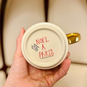 May include: A cream-colored ceramic mug with a gold handle. The bottom of the mug features red text that reads "NO&Euml;L &Agrave; PARIS" and "by ANTHROPOLOGIE". The mug is held in a person's hand.