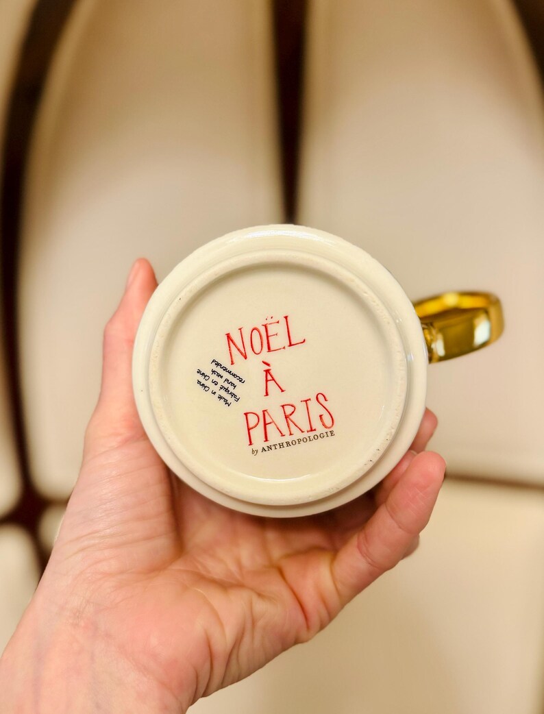 May include: A cream-colored ceramic mug with a gold handle. The bottom of the mug features red text that reads "NO&Euml;L &Agrave; PARIS" and "by ANTHROPOLOGIE". The mug is held in a person's hand.