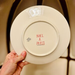 May include: A cream-colored ceramic plate with the words "NOEL &Agrave; PARIS" in red. The plate has a circular design and is held by a hand. The plate is from Anthropologie.