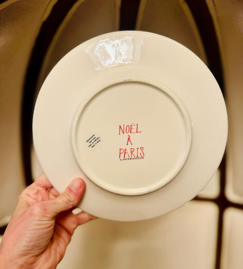 May include: A cream-colored ceramic plate with the words "NOEL &Agrave; PARIS" in red. The plate has a circular design and is held by a hand. The plate is from Anthropologie.