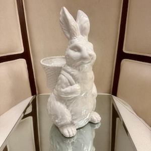 Può includere: Statuetta di coniglio in ceramica bianca con un cestino sulla schiena. Il coniglio è posizionato su una superficie a specchio, riflettendo la sua forma. La statuetta ha dettagli ed è un oggetto decorativo.