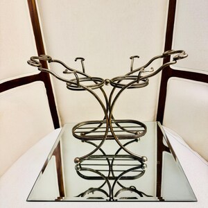 May include: A metallic bronze-toned wire rack featuring two circular holders connected by curved supports. The structure includes a rectangular base with horizontal wire slats and decorative scrollwork at the top edges of the frame.