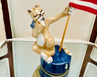 Può includere: Una statuetta di gatto in ceramica con pelo color marrone chiaro e petto bianco, seduta su una base rotonda blu. Il gatto tiene un'asta di legno sormontata da una bandiera americana in miniatura con strisce rosse e bianche e un campo blu con stelle.