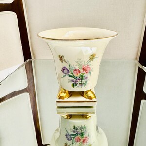 May include: A cream-colored ceramic vase with a flared rim and gold-painted trim. The body features a floral bouquet of pink, purple, and yellow flowers. The vase is supported by a square base with four small, gold-painted feet at the bottom.