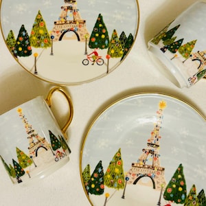 May include: A set of two plates and two mugs with a winter holiday theme. The light blue plates and mugs feature a gold rim and a design of the Eiffel Tower decorated with lights, Christmas trees, and a cyclist. Snowflakes are scattered across the background.