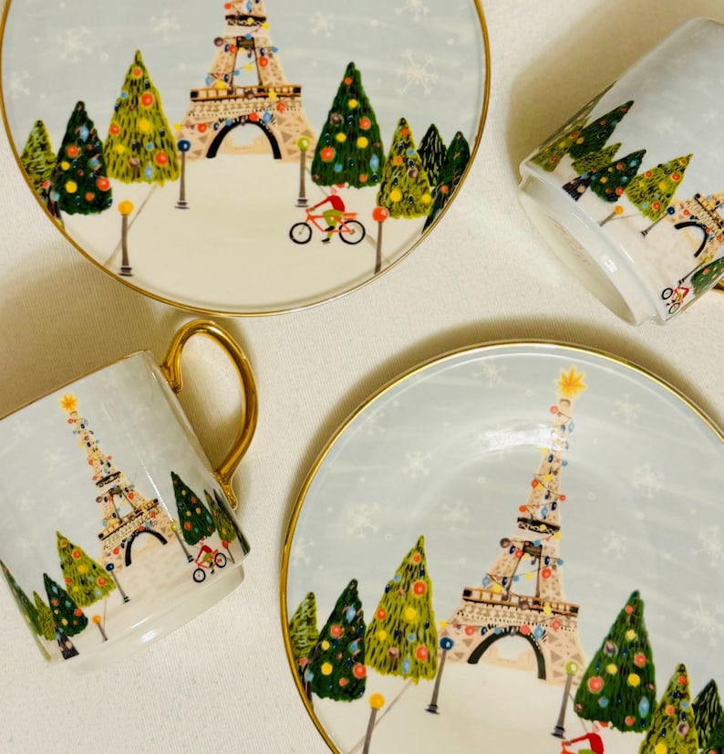 May include: A set of two plates and two mugs with a winter holiday theme. The light blue plates and mugs feature a gold rim and a design of the Eiffel Tower decorated with lights, Christmas trees, and a cyclist. Snowflakes are scattered across the background.