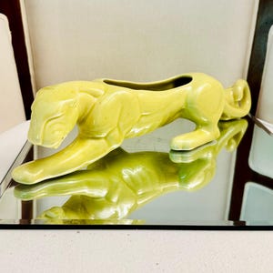 May include: A chartreuse ceramic planter shaped like a prowling panther. The feline figure is depicted in a low, stalking stance with its head lowered and tail curled. The glossy, smooth finish reflects light, highlighting the sculpted muscular form.