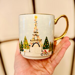 May include: A white ceramic mug with a gold handle and rim. The mug features a colorful illustration of the Eiffel Tower decorated with lights, surrounded by Christmas trees and a cyclist. The design is set against a light blue background.