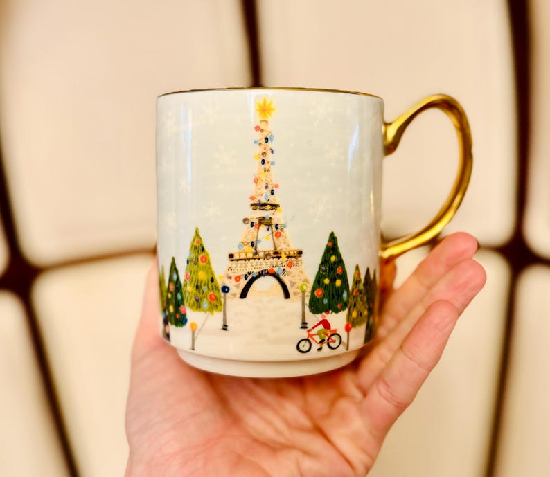 May include: A white ceramic mug with a gold handle and rim. The mug features a colorful illustration of the Eiffel Tower decorated with lights, surrounded by Christmas trees and a cyclist. The design is set against a light blue background.