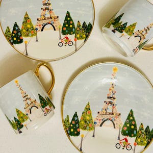 May include: Four pieces of holiday-themed tableware: two plates and two mugs. Each piece features a light blue background with a festive scene of the Eiffel Tower decorated with lights, Christmas trees, and a cyclist. The rims and handles are gold.