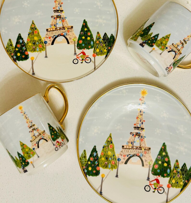 May include: Four pieces of holiday-themed tableware: two plates and two mugs. Each piece features a light blue background with a festive scene of the Eiffel Tower decorated with lights, Christmas trees, and a cyclist. The rims and handles are gold.