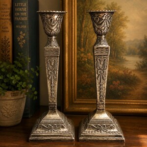 May include: A pair of silver-toned metal candlesticks featuring intricate embossed details, including ram heads, floral swags, and ribbon motifs. Each candlestick has a square base, a tapered stem, and a wide, scalloped rim at the top.