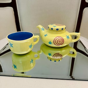 以下が含まれることがあります： 青い星と渦巻き模様の黄色いセラミックティーポットとティーカップのセット。ティーカップは内側が青色です。ティーポットには渦巻き模様の蓋が付いています。セットは反射面の上にあります。