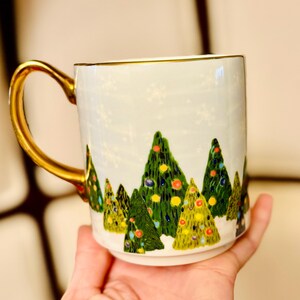 May include: White ceramic mug with a gold handle and rim, decorated with a winter forest scene of green Christmas trees adorned with colorful ornaments. The mug has a light blue background with white snowflakes.