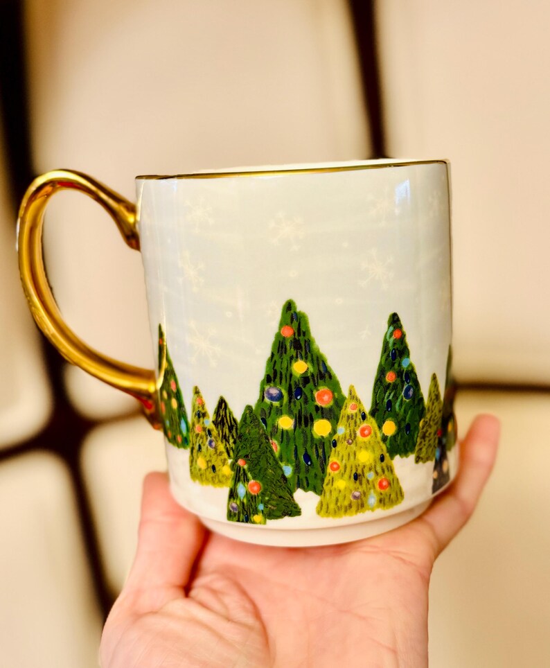 May include: White ceramic mug with a gold handle and rim, decorated with a winter forest scene of green Christmas trees adorned with colorful ornaments. The mug has a light blue background with white snowflakes.