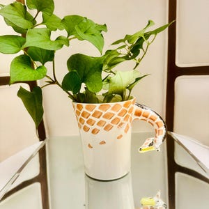 Peut inclure: Une tasse en céramique avec un corps crème orné d'un motif de grille marron rappelant une girafe. L'anse est façonnée en forme de cou et de tête de girafe, avec des traits du visage peints. Une plante verte feuillue est placée dans la tasse.