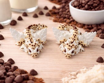 May include: Two dragon-shaped pastries with white wings and tan spots are displayed on a wooden surface, surrounded by chocolate chips, cocoa powder, and a bowl of chocolate chips. Two glasses of a white liquid are in the background.