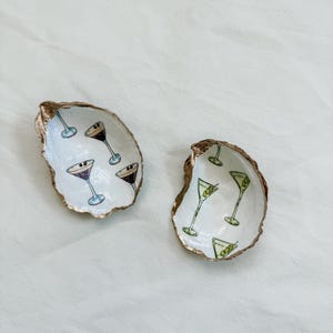 May include: Two oyster shell dishes with gold rims. One shell has martini glasses with purple and black drinks, the other has green olive martinis. The shells are on a white surface.
