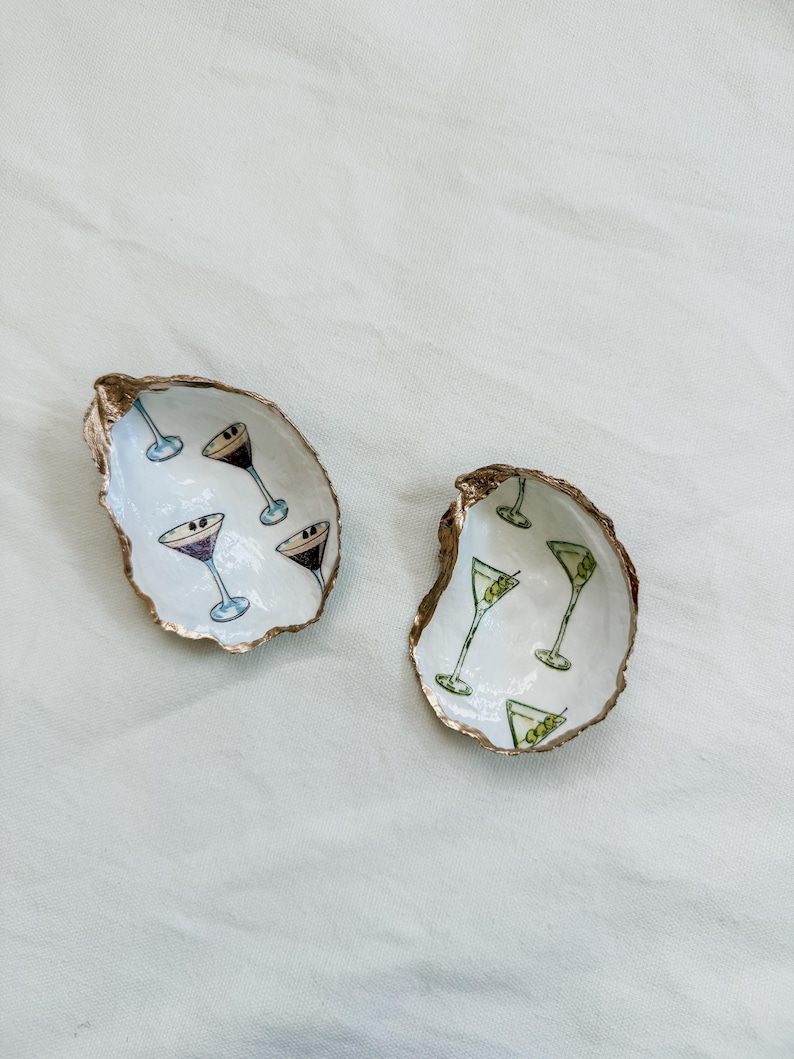 May include: Two oyster shell dishes with gold rims. One shell has martini glasses with purple and black drinks, the other has green olive martinis. The shells are on a white surface.