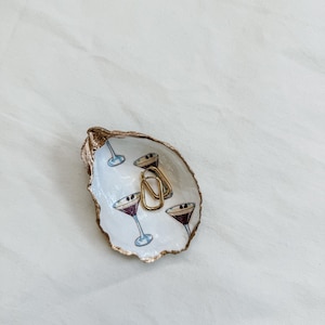 May include: An oyster shell trinket dish with a gold rim and a painted interior featuring martini glasses. Two gold hoop earrings rest inside the dish. The dish is white with blue and purple martini glasses.