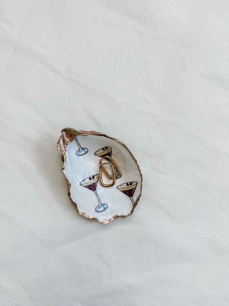 May include: An oyster shell trinket dish with a gold rim and a painted interior featuring martini glasses. Two gold hoop earrings rest inside the dish. The dish is white with blue and purple martini glasses.