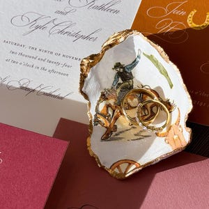 May include: Wedding invitation suite featuring a decorative oyster shell ring dish with gold trim and a cowboy illustration. Gold wedding rings rest inside the dish. Invitations are in shades of white, orange, and burgundy.