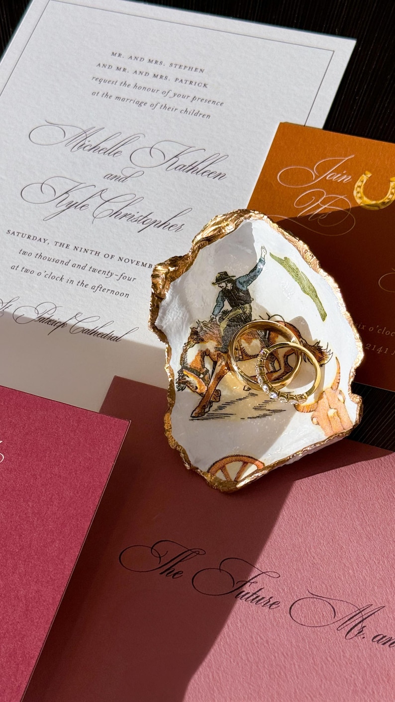 May include: Wedding invitation suite featuring a decorative oyster shell ring dish with gold trim and a cowboy illustration. Gold wedding rings rest inside the dish. Invitations are in shades of white, orange, and burgundy.