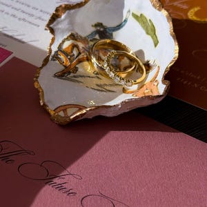 May include: A decorative, white, shell-shaped dish with a gold rim holds two gold wedding rings. The dish features a painted scene of a cowboy on a horse. Wedding invitations in the background.