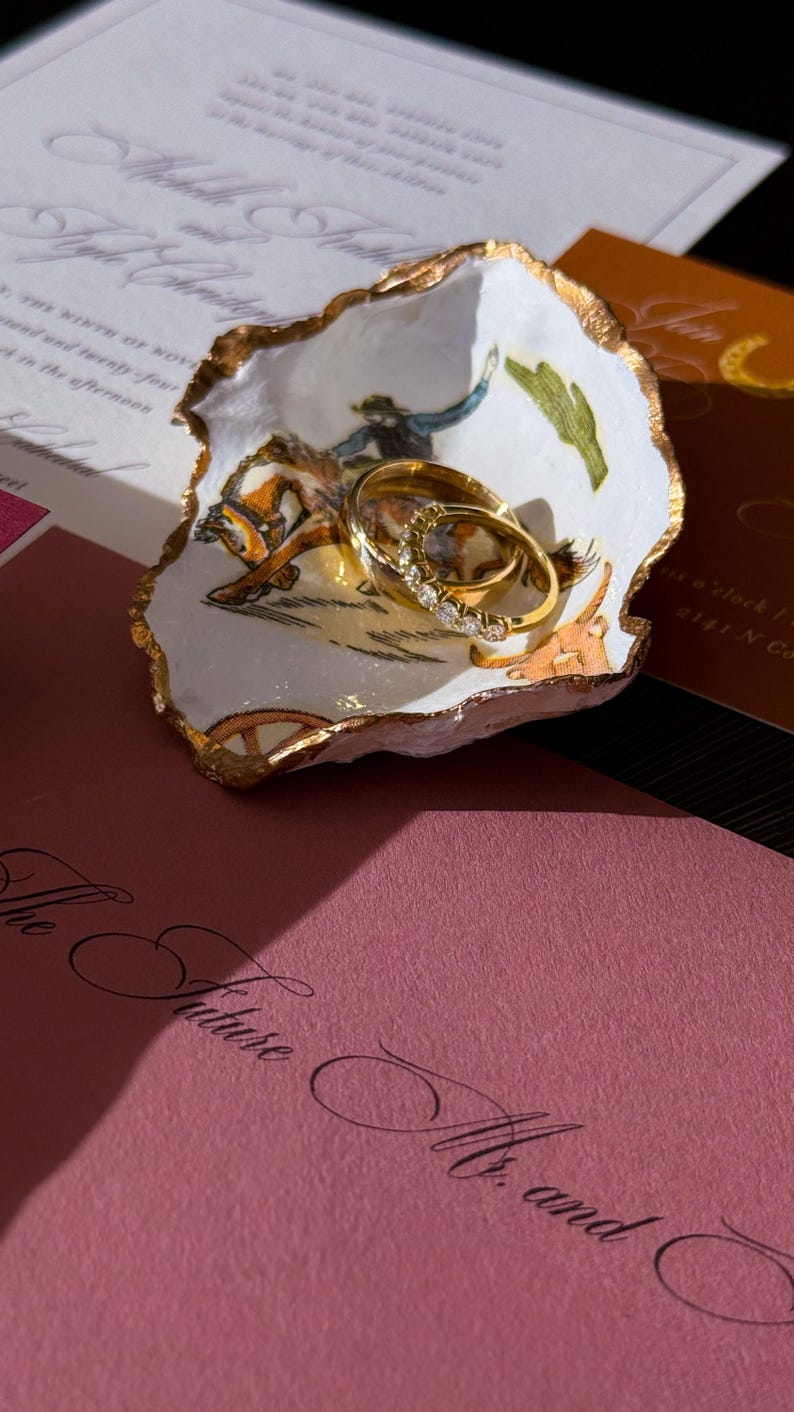 May include: A decorative, white, shell-shaped dish with a gold rim holds two gold wedding rings. The dish features a painted scene of a cowboy on a horse. Wedding invitations in the background.