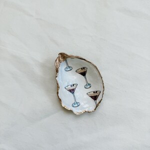May include: A decorative oyster shell dish with a gold-painted rim. The interior is white and features hand-painted martini glasses. The glasses have a purple liquid and a black olive. A unique and stylish home decor item.