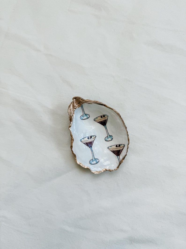 May include: A decorative oyster shell dish with a gold-painted rim. The interior is white and features hand-painted martini glasses. The glasses have a purple liquid and a black olive. A unique and stylish home decor item.