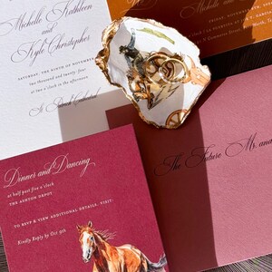 May include: Wedding invitation suite featuring a watercolor horse illustration, gold rings in a decorative dish, and various invitation cards with elegant calligraphy. The color palette includes burgundy, gold, and cream.