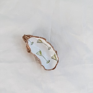 May include: An oyster shell dish with a white interior featuring a martini design. The martini glasses are green and black, with olive details. The shell has a natural, textured, brown exterior.