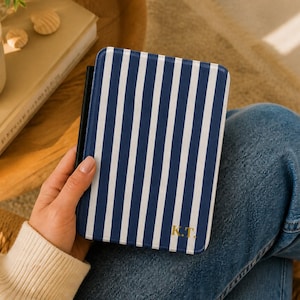 Könnte beinhalten: Eine Tablet-Hülle mit marineblauen und weißen vertikalen Streifen und goldenen Initialen "K.T." wird gehalten. Im Hintergrund sind ein Buch und Muscheln zu sehen.