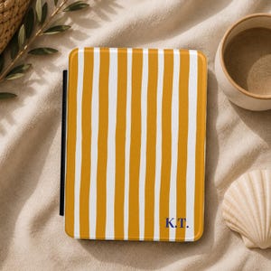 Könnte beinhalten: Ein gelb-weiß gestreifter Buchumschlag mit den Initialen "K.T." in Marineblau. Der Umschlag ist rechteckig und hat einen schwarzen Rücken. Der Hintergrund ist ein beiger Stoff mit einer Muschel und einer kleinen Keramiktasse.