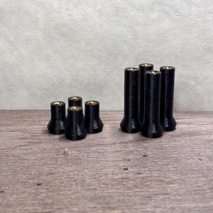 May include: Several black, cylindrical objects with a flared base and a gold-coloured interior are arranged on a wooden surface. The objects are small, possibly decorative or functional items, and are set against a neutral background.