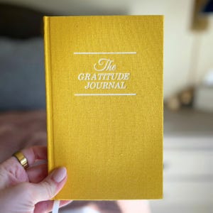 以下が含まれることがあります： 「The GRATITUDE JOURNAL」の白い文字が入ったマスタードイエローのジャーナル。 金色の指輪をした手でジャーナルが持たれています。 ジャーナルはテクスチャード加工されたカバーと白いリボンブックマークが付いています。