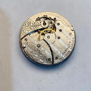 May include: Close-up of the inner workings of a silver pocket watch. The watch face is engraved with "American Waltham Watch Co.", "Barrel 18 Size 11", and "Safety".