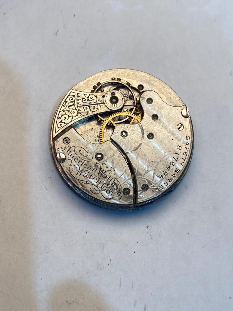 May include: Close-up of the inner workings of a silver pocket watch. The watch face is engraved with "American Waltham Watch Co.", "Barrel 18 Size 11", and "Safety".