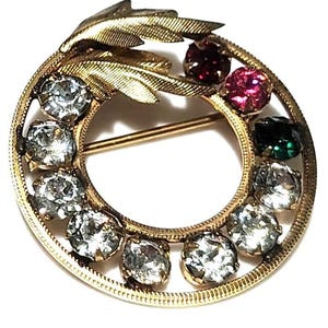 May include: A circular gold-tone brooch featuring a textured leaf design at the top. The frame is set with a series of round faceted stones in clear, dark red, pink, and dark green colors, arranged along the inner edge of the circular metal band.