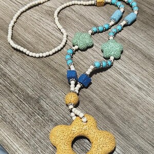May include: A beaded necklace featuring a large yellow porous flower-shaped pendant, small white round beads, and various textured lava stone beads in blue, light blue, green, and yellow, accented with small silver-toned metallic spacer beads.