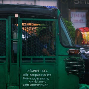 Könnte beinhalten: Ein grüner Auto-Rikscha mit einem Metallkäfig und einem Schild mit der Aufschrift "অভিযোগ-৯৯৯" (Abhijoga-999) mit Kontaktinformationen. Der Rikscha steht auf einer Straße mit anderen Fahrzeugen im Hintergrund.
